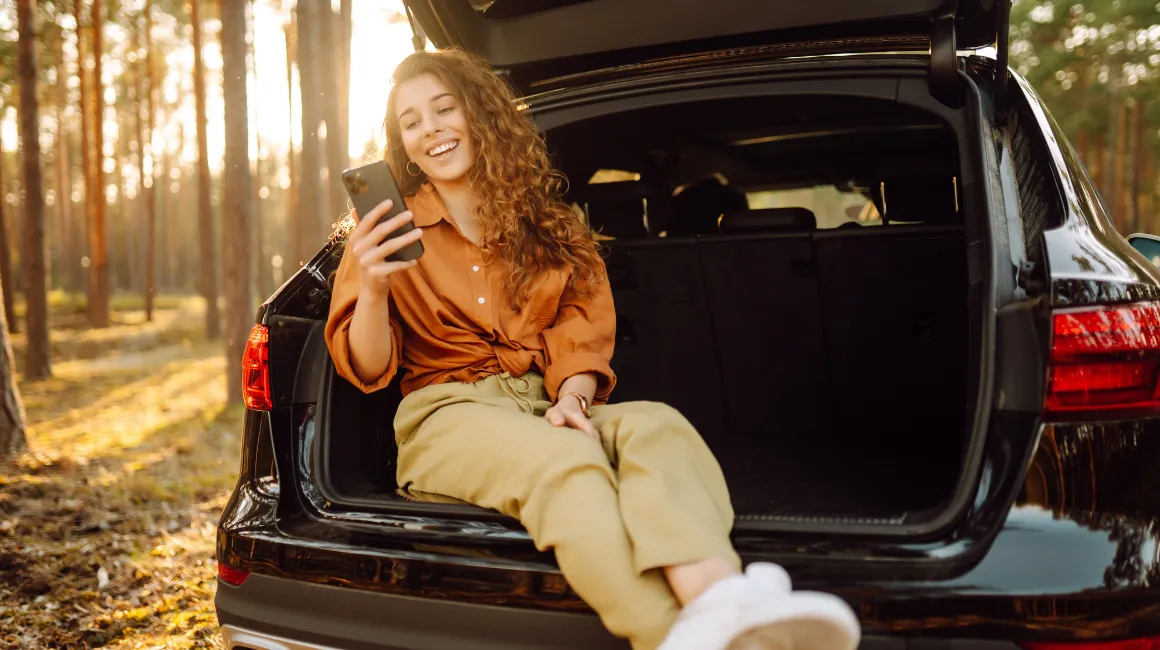 Zadowolona młoda kobieta sprawdza coś na smartfonie, siedząc w otwartym bagażniku samochodu