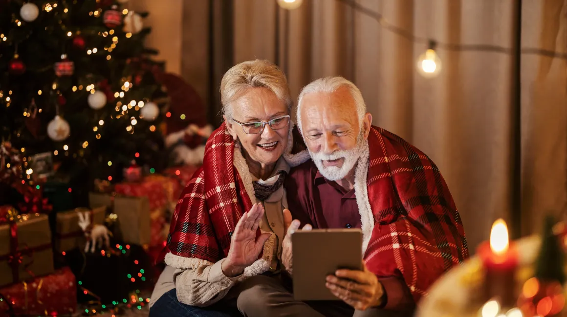 Uśmiechnięci seniorzy pod kocem w kratę przy choince rozmawiają przez tablet w świątecznej atmosferze